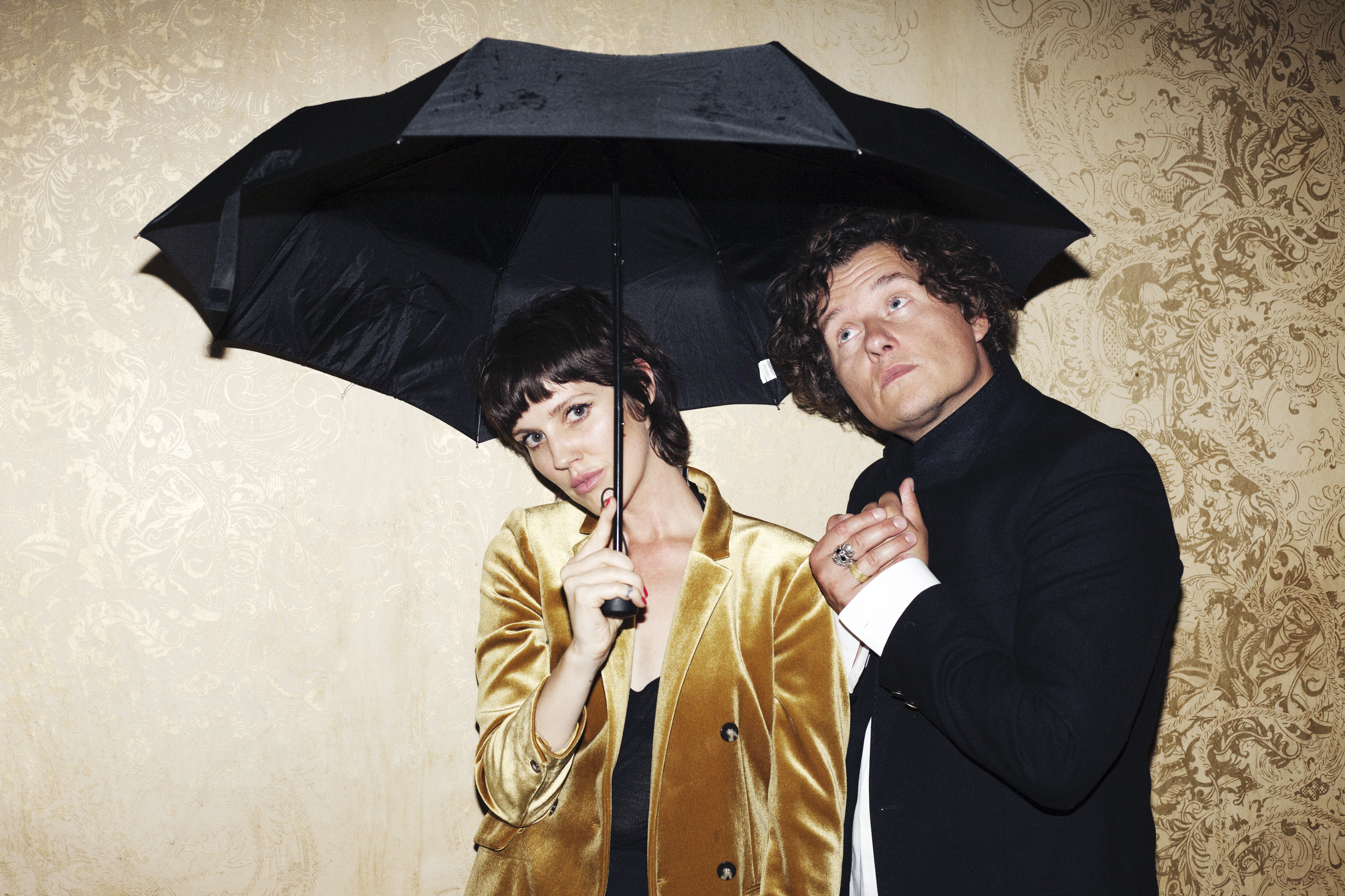 The music duo Rhonda Star consists of Ronja Violett and her boyfriend Alex Voigt. Together they stand under an umbrella, which looks funny because there is wallpaper in the background. Ronja holds the umbrella and looks into the camera with her head tilted. Alex has turned his gaze towards the sky.